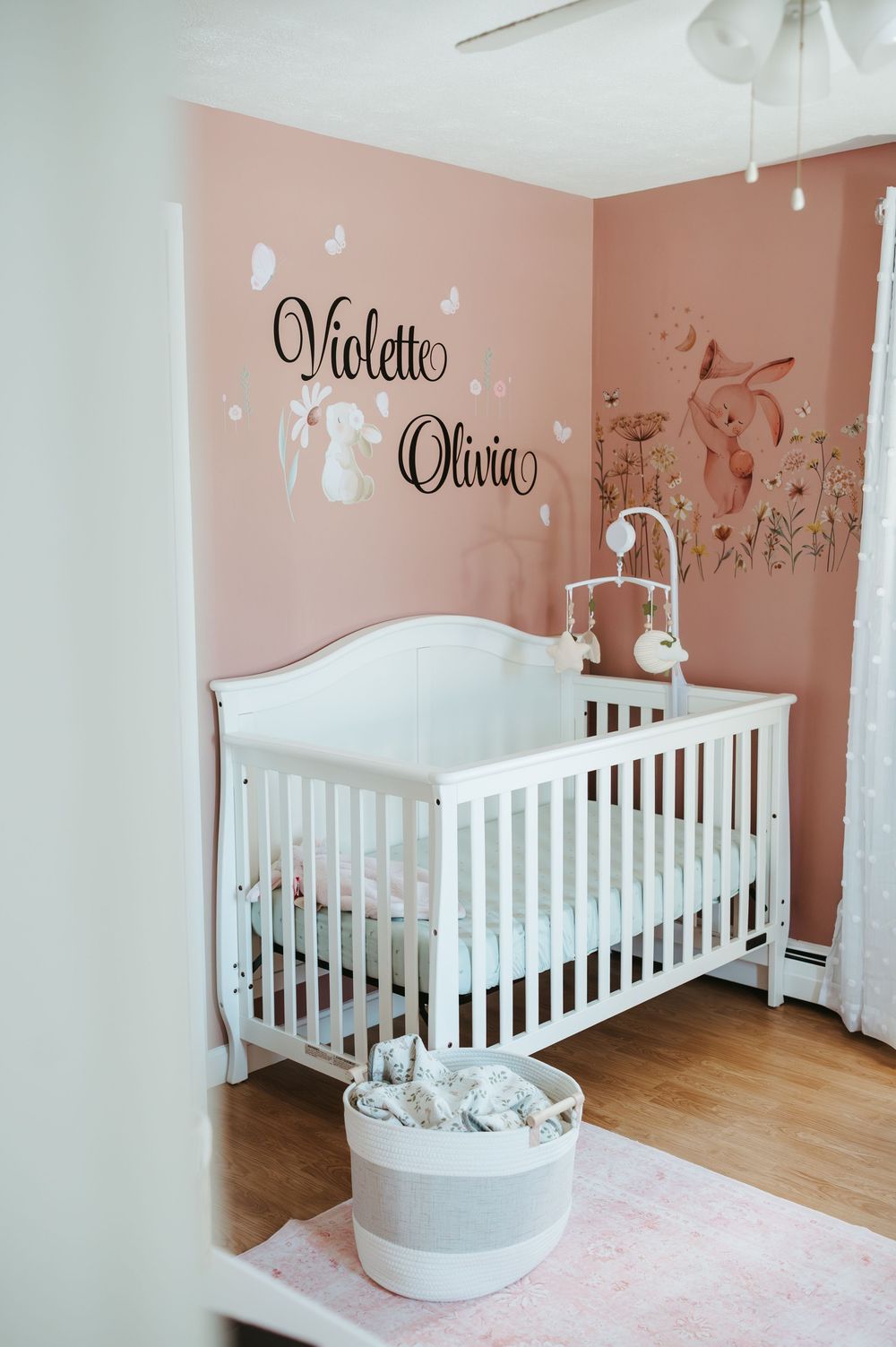 Pink nursery with white crib, Violette Olivia wall decal, floral fox mural, and soft pink rug on hardwood floors.