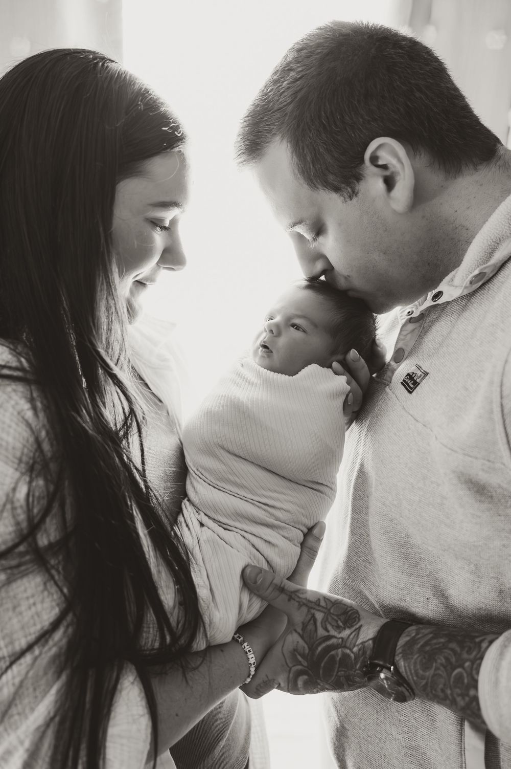 Black and white photo of new parents lovingly holding their swaddled newborn baby, father kissing infant's head.