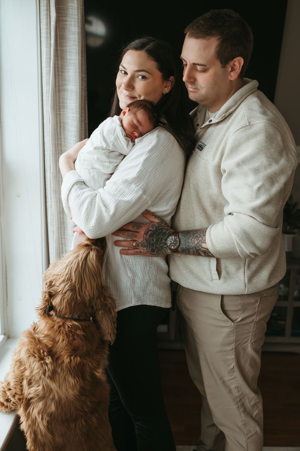 A young couple holds their newborn baby by a window, with a fluffy golden dog sitting beside them.