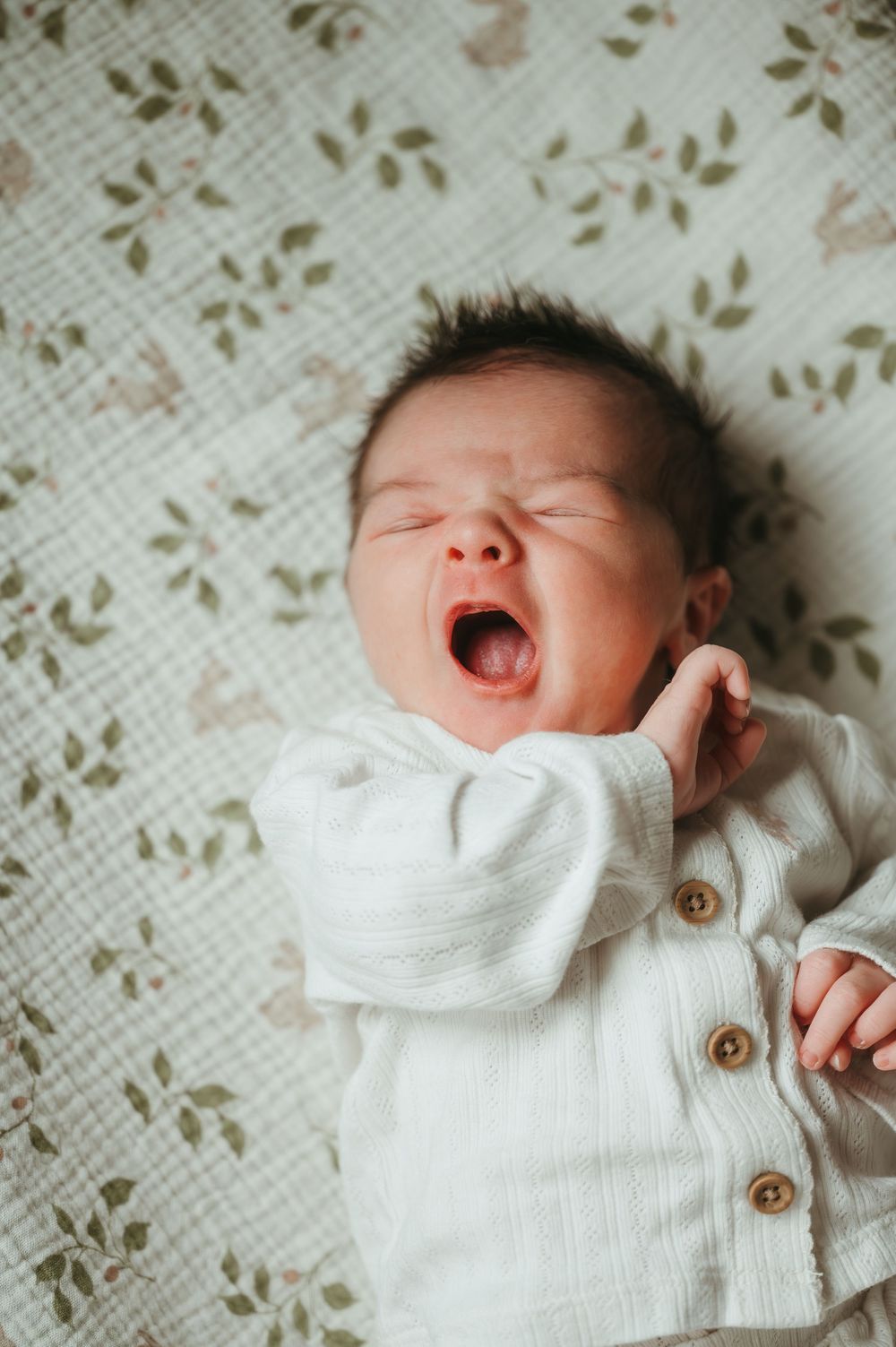 A newborn baby yawning while lying on a floral blanket, dressed in a white cotton button-up outfit.