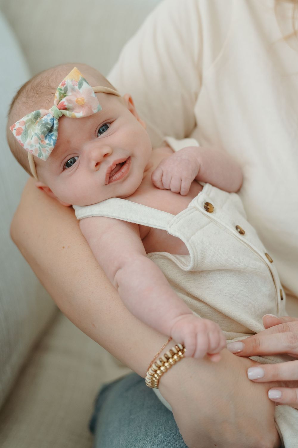 A smiling baby wearing a floral bow headband and white romper rests on someone's holding arms.