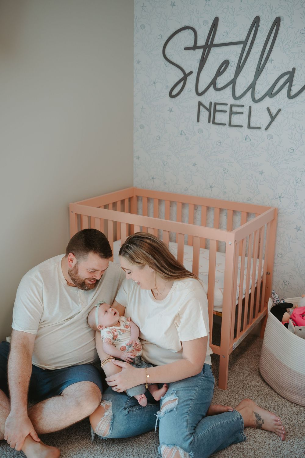 Parents sit with their newborn baby in a nursery room decorated with the name Stella Neely on the wall and a pink crib.