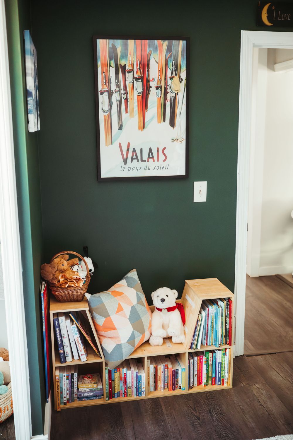 Children's reading nook with wooden bookshelf, stuffed bear, geometric pillow, and Valais ski poster on dark green wall.