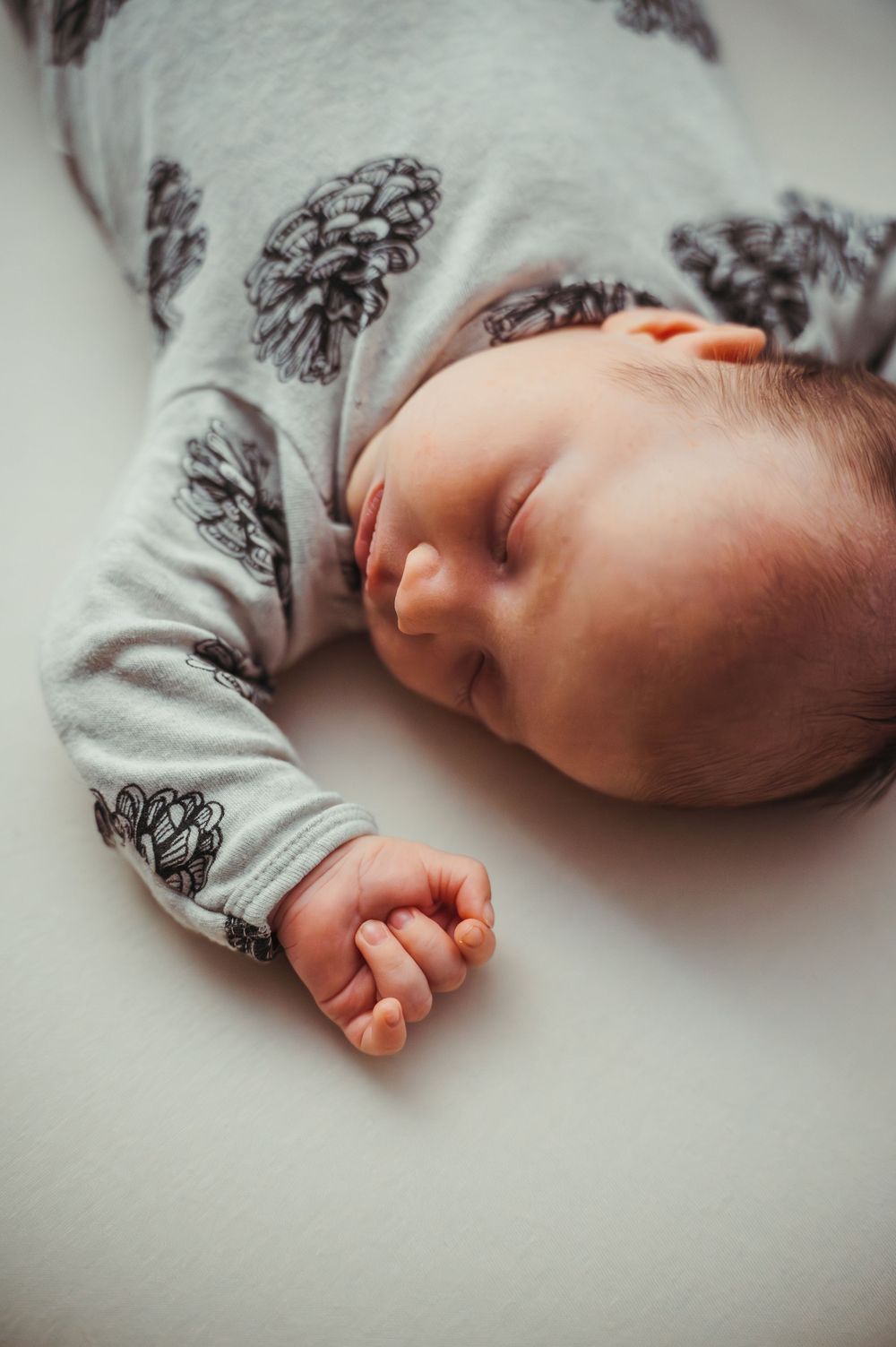 Newborn baby sleeping peacefully in a gray pine cone print onesie, lying on a white surface.