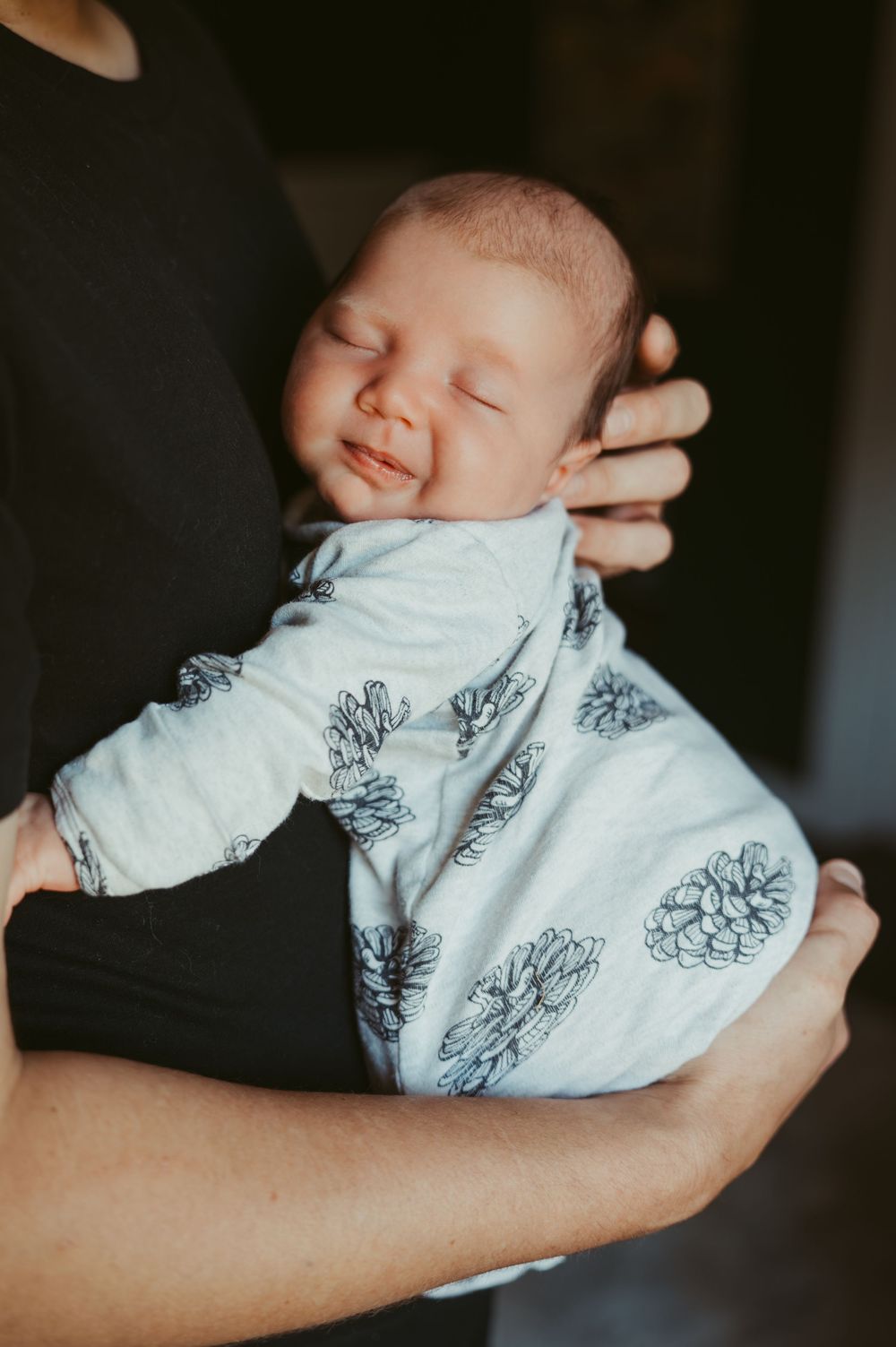 A newborn baby sleeping peacefully in a pine cone patterned onesie, cradled in an adult's arms.