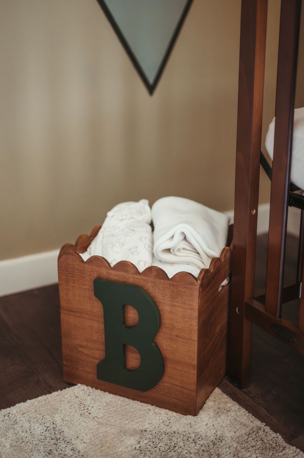 Wooden scalloped box with green letter B holding folded white blankets, beside a dark wood crib in a nursery.
