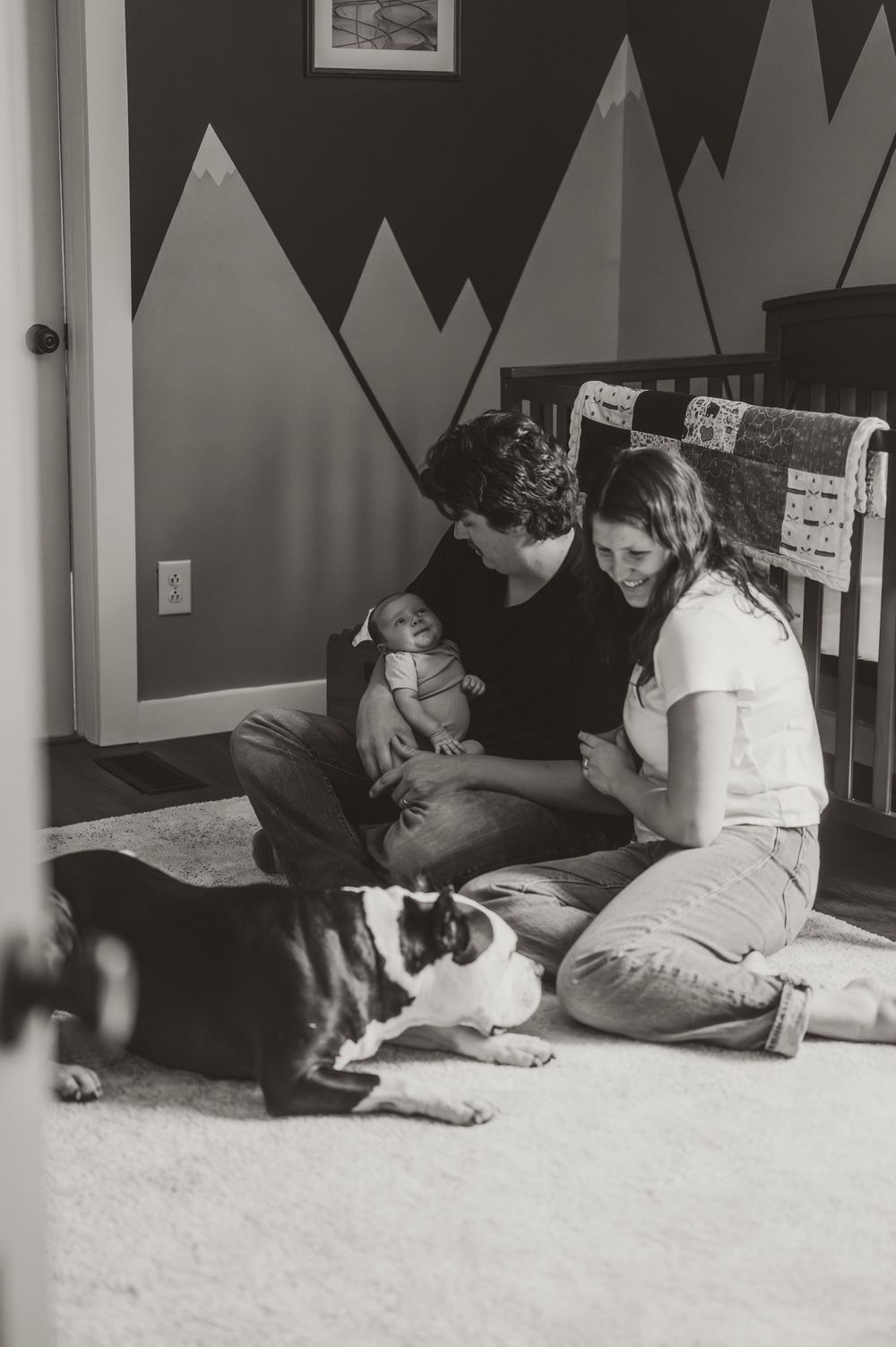 Black and white photo of parents sitting on floor with newborn baby and Boston Terrier dog in mountain-themed nursery.