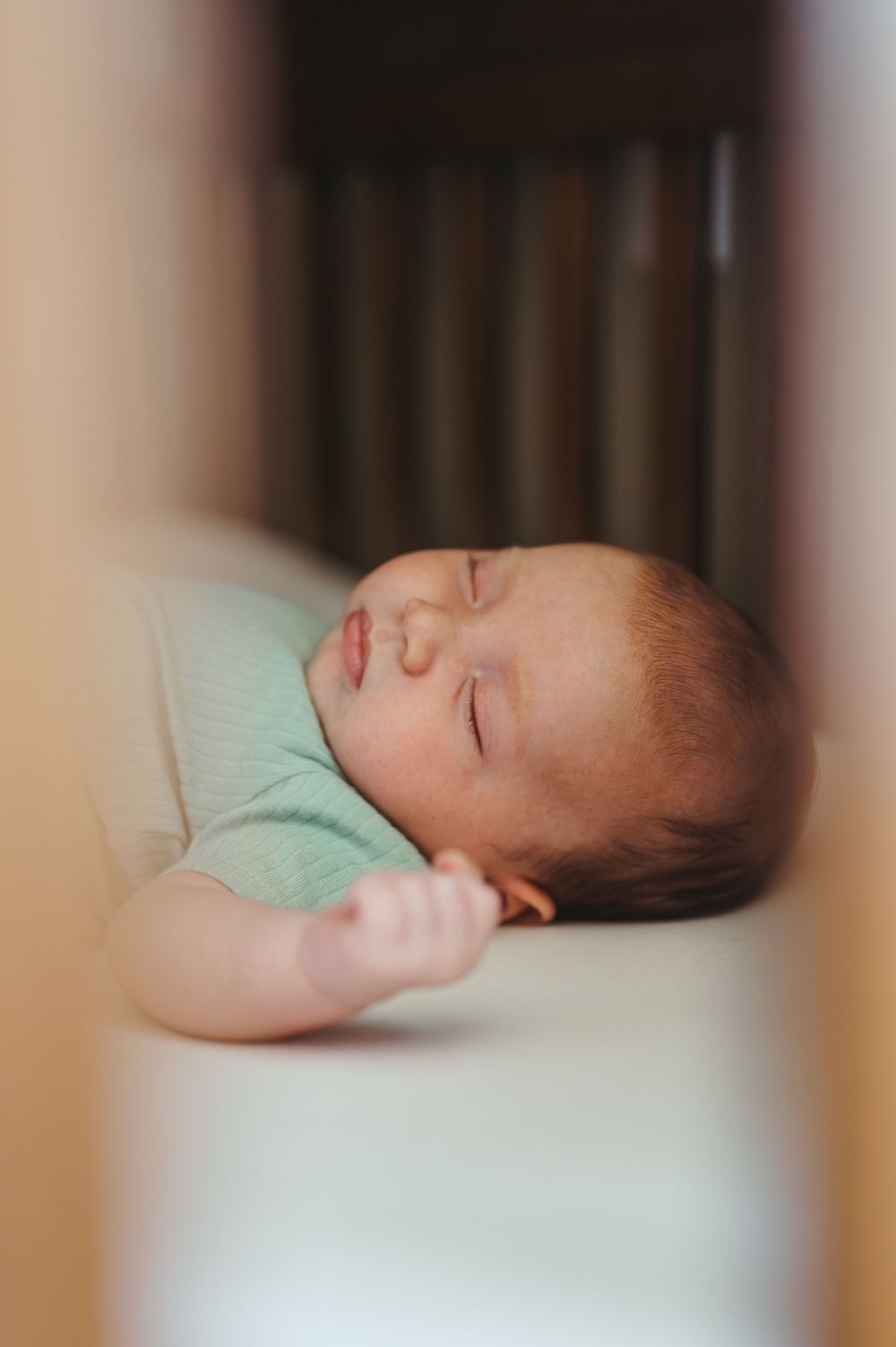 A peaceful newborn baby sleeping soundly in a white crib, wearing a mint green onesie with tiny fist visible.