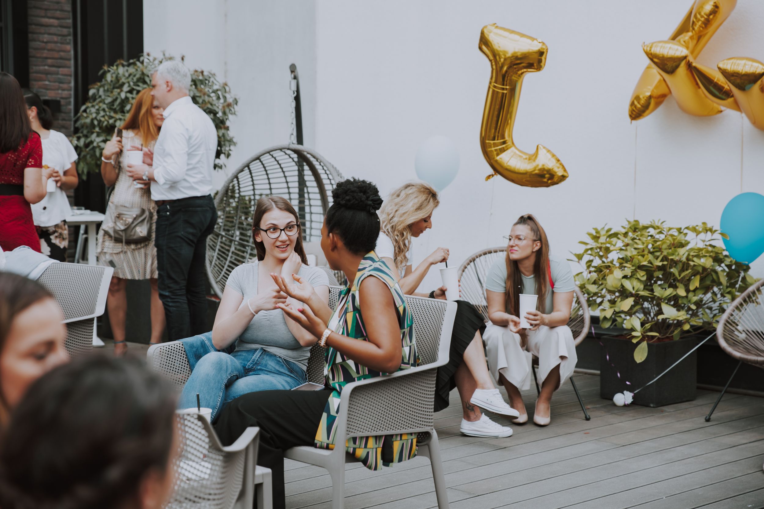 Outdoor party setting with gold balloon decorations and guests seated in chairs.