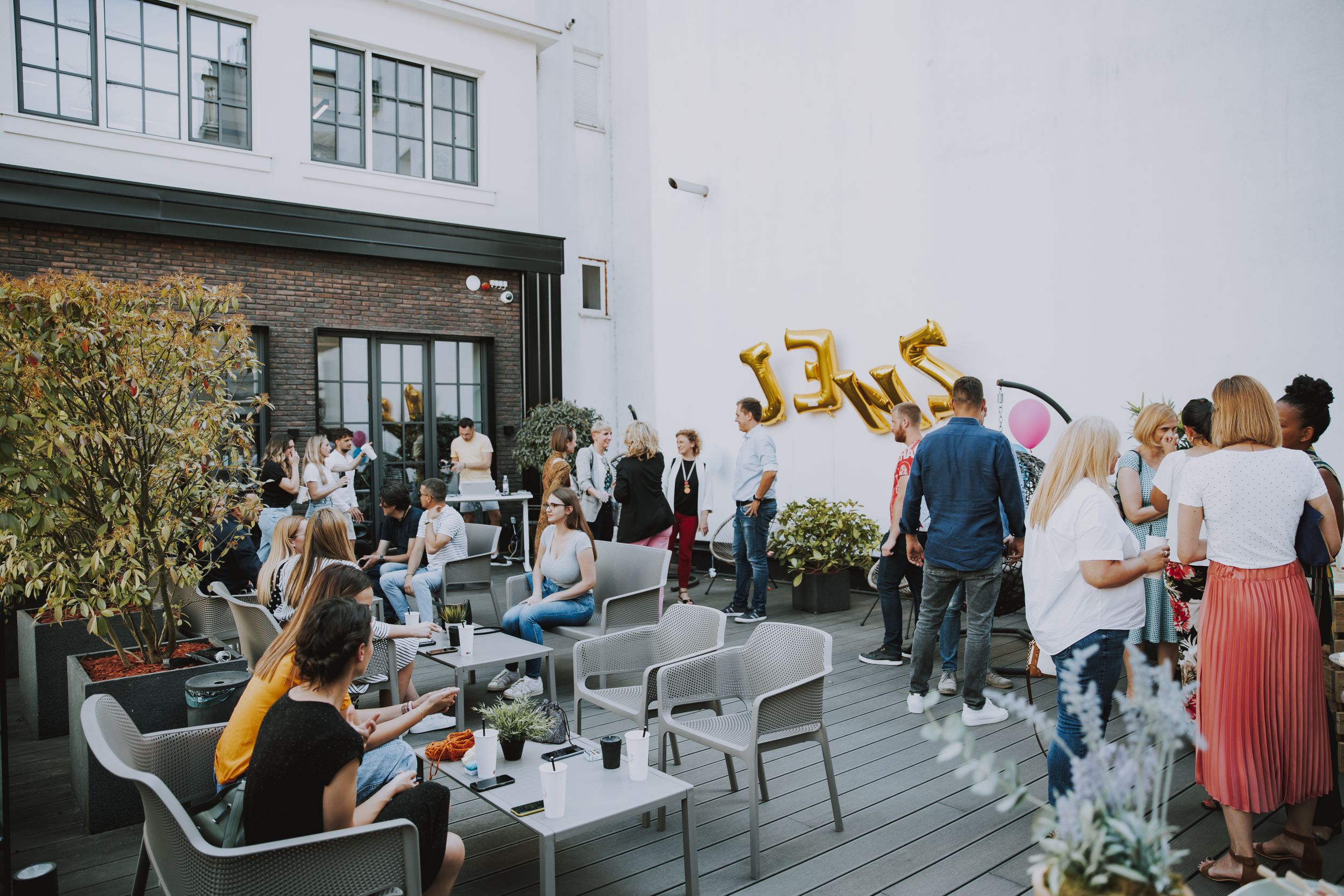 Outdoor rooftop party setup with chairs and gold number balloons against white building.