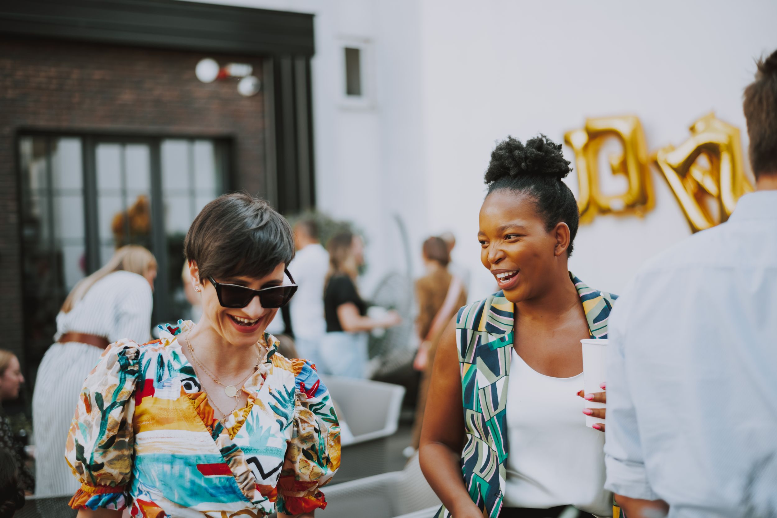 Two friends in colorful summer outfits sharing a laugh at an outdoor social gathering.