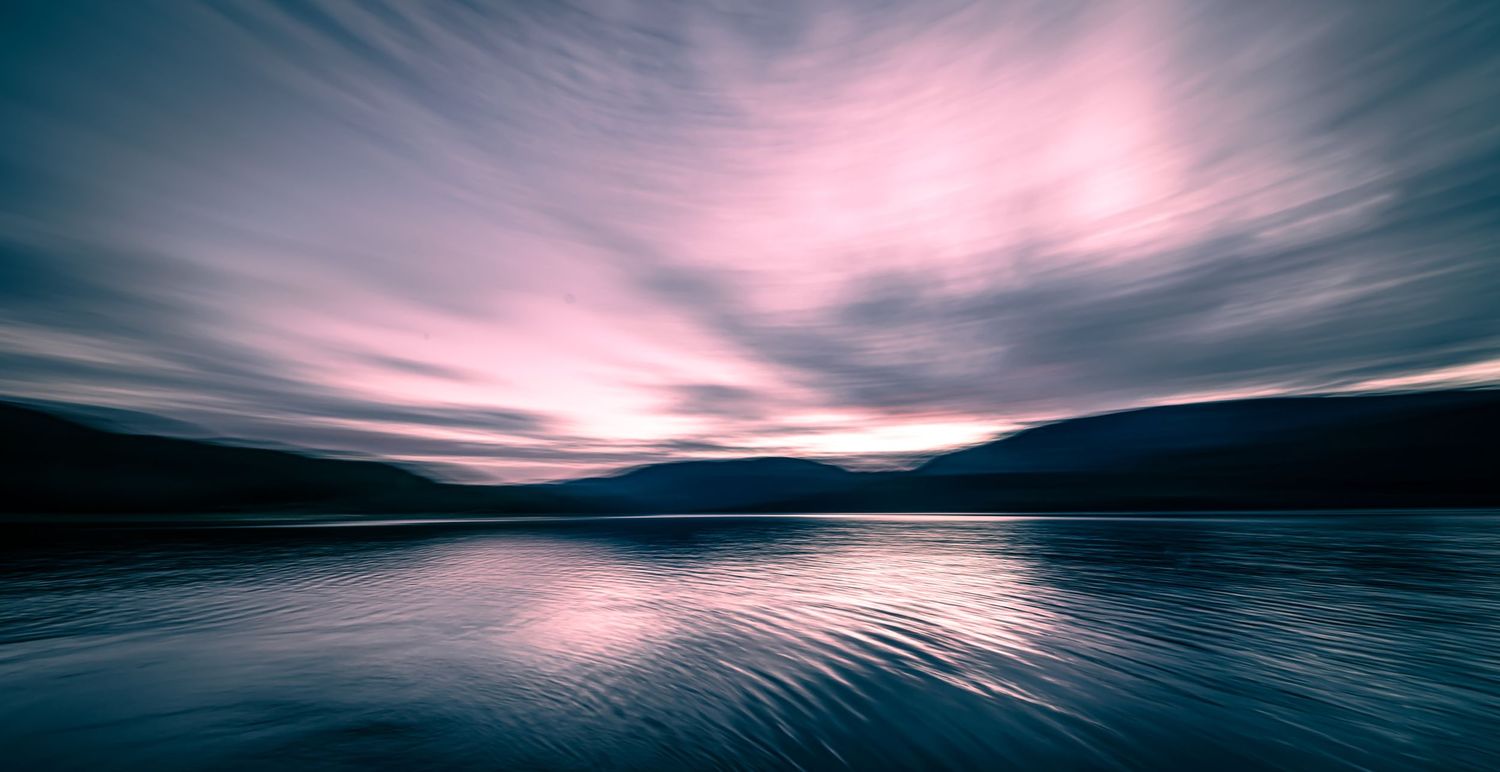 Dramatic pink and purple clouds reflect on a tranquil lake at sunset with mountains silhouetted in the distance.