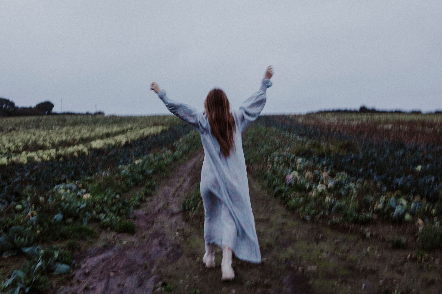 Figure in white dress raises arms joyfully while walking down path between flowering fields.