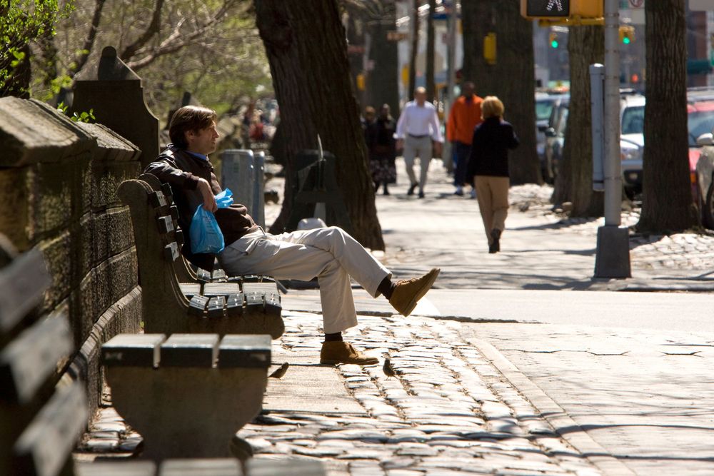 A person relaxes on a park bench on a sunny sidewalk surrounded by trees and pedestrians.