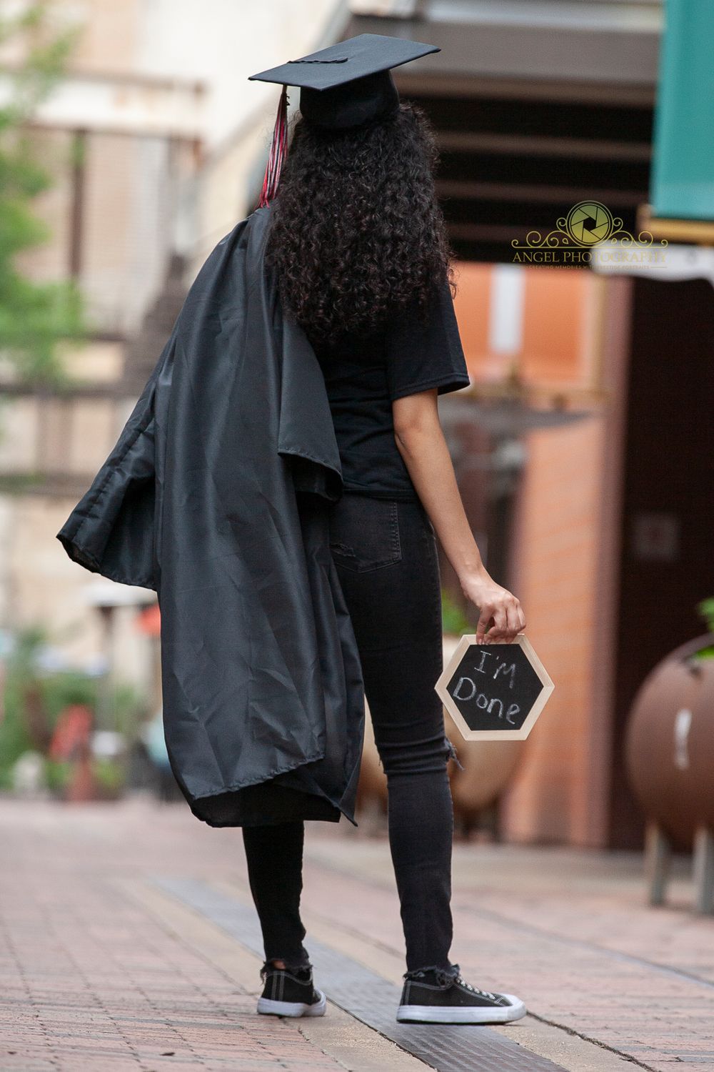 Graduate in black cap and gown holds diploma while walking on brick path.