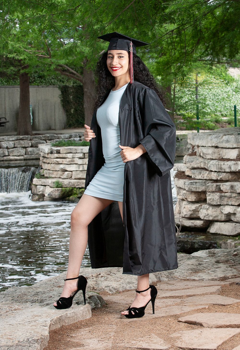 Graduate in cap and gown poses by stone waterfall feature in outdoor setting.