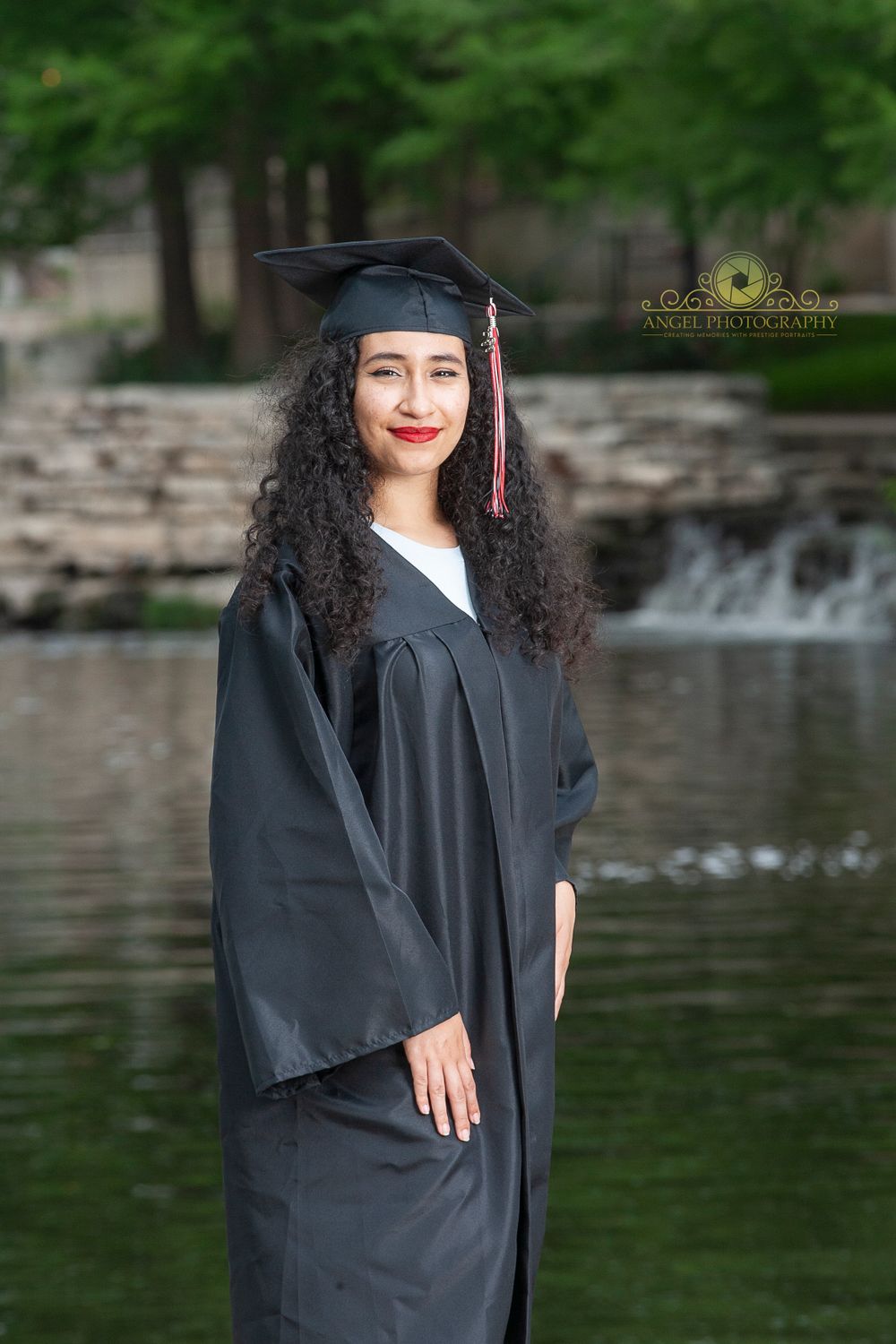 Graduate in traditional black cap and gown poses by scenic water feature.