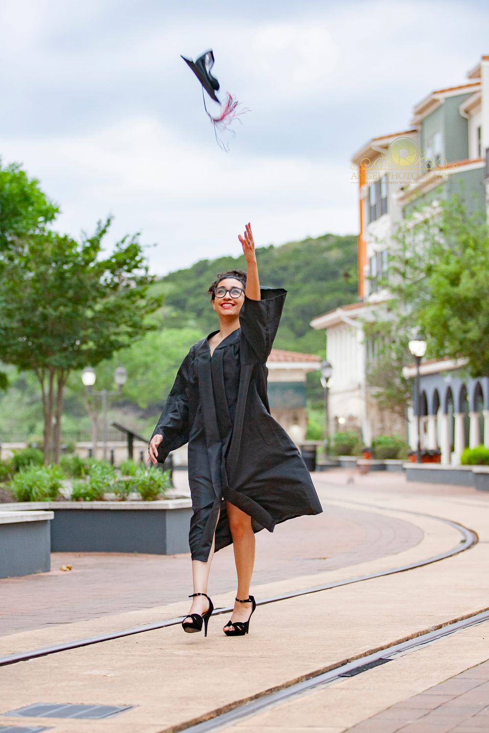 Graduate throws cap in celebration on curved walkway with buildings in background.