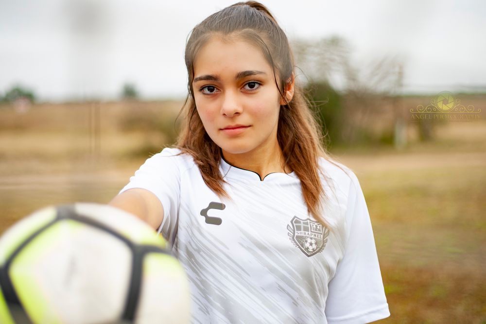 Soccer player in white jersey poses with ball on field during golden hour.