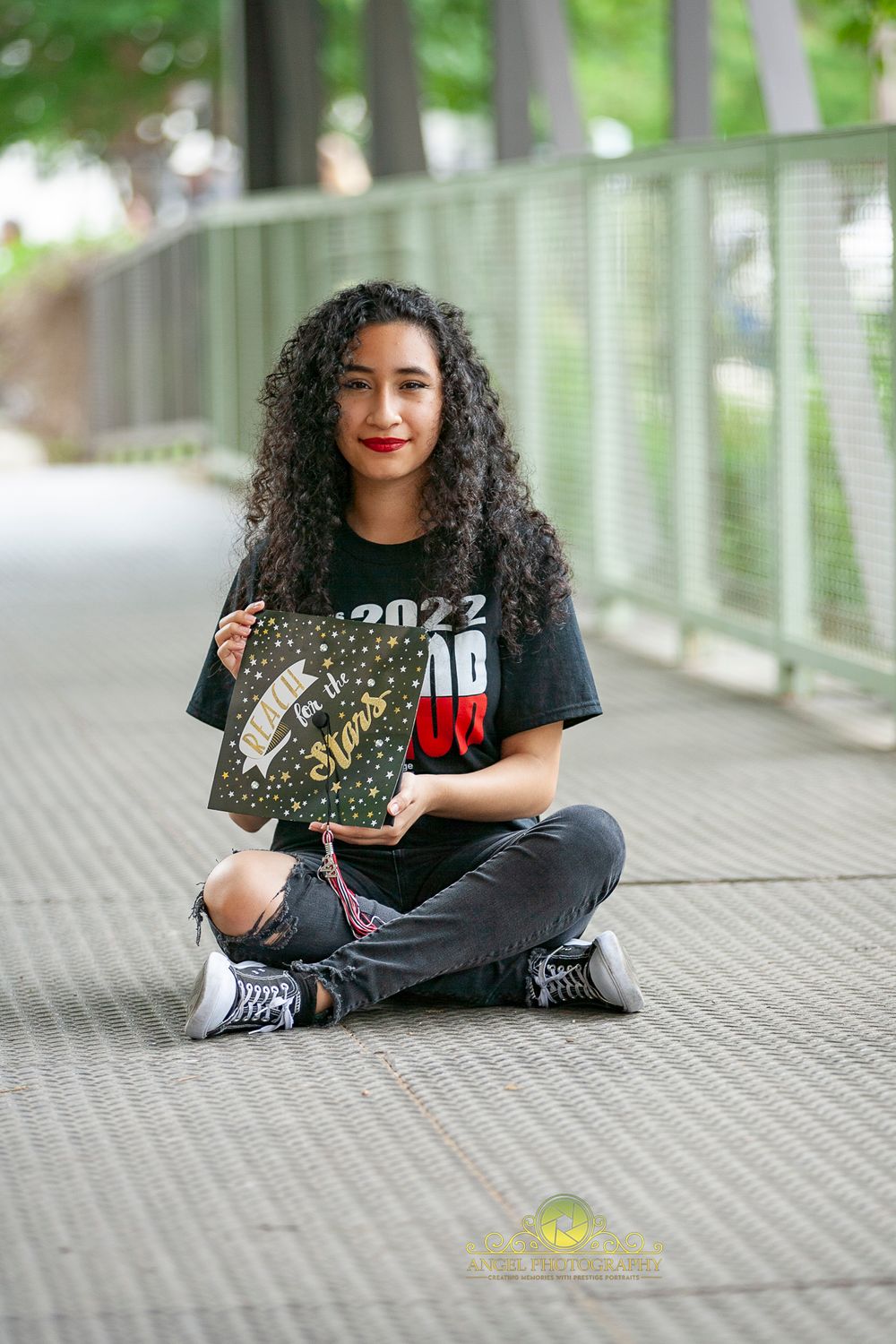 A graduate in casual attire sits cross-legged on a covered walkway holding their diploma.
