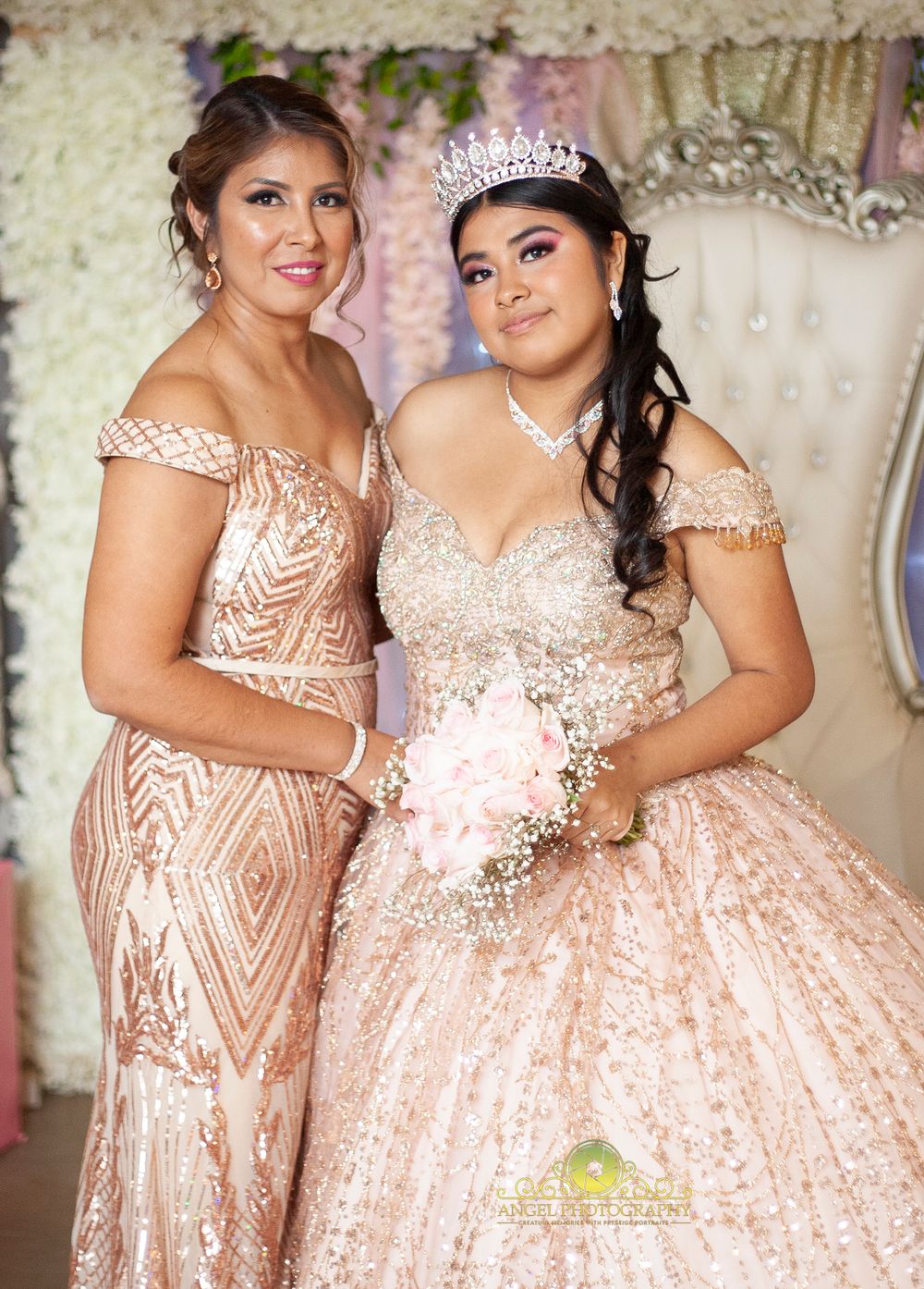 Two women in sparkly formal gowns pose at a quinceañera celebration with floral backdrop.