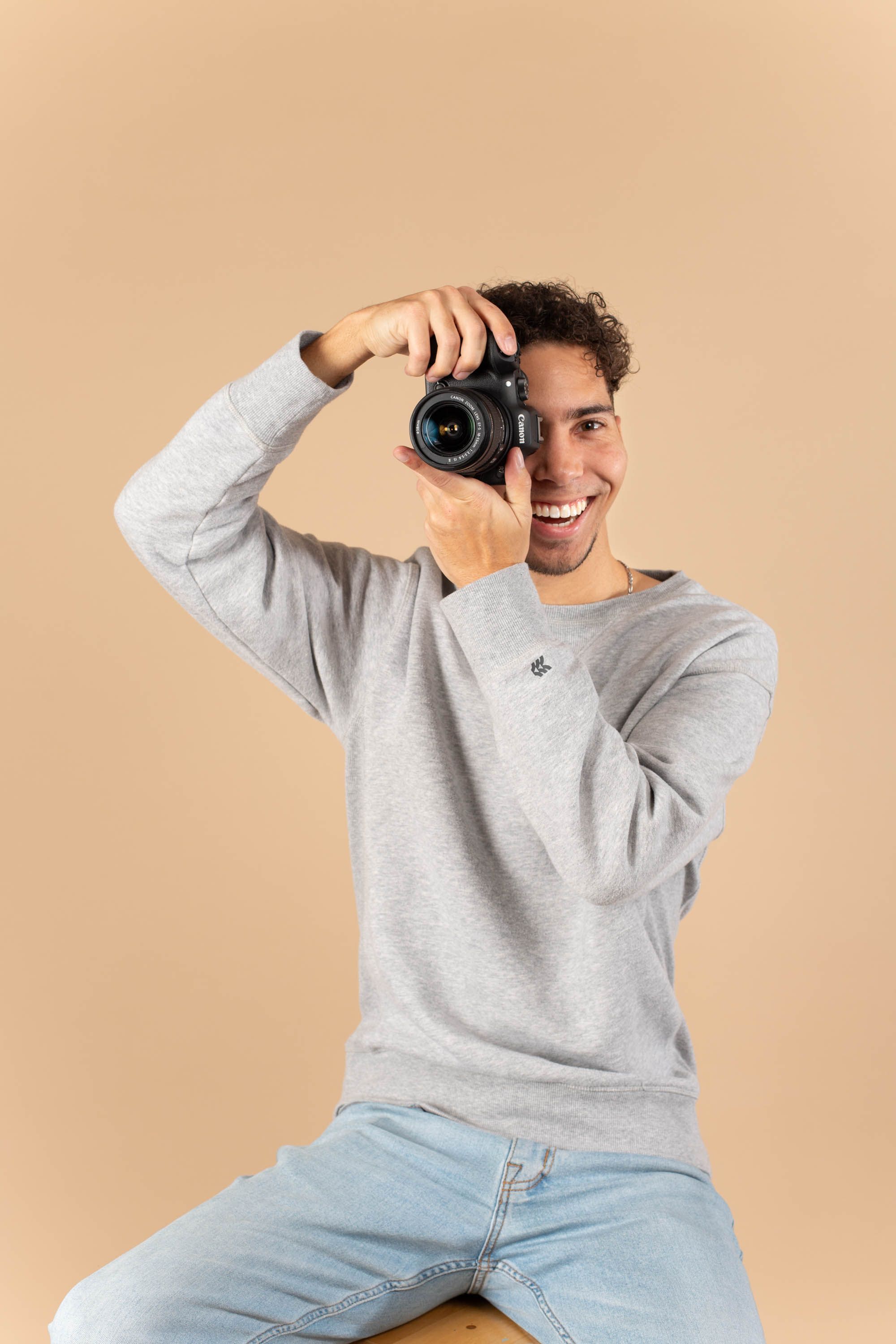 Happy male photographer raising a DSLR camera to his face while seated on a stool in studio.