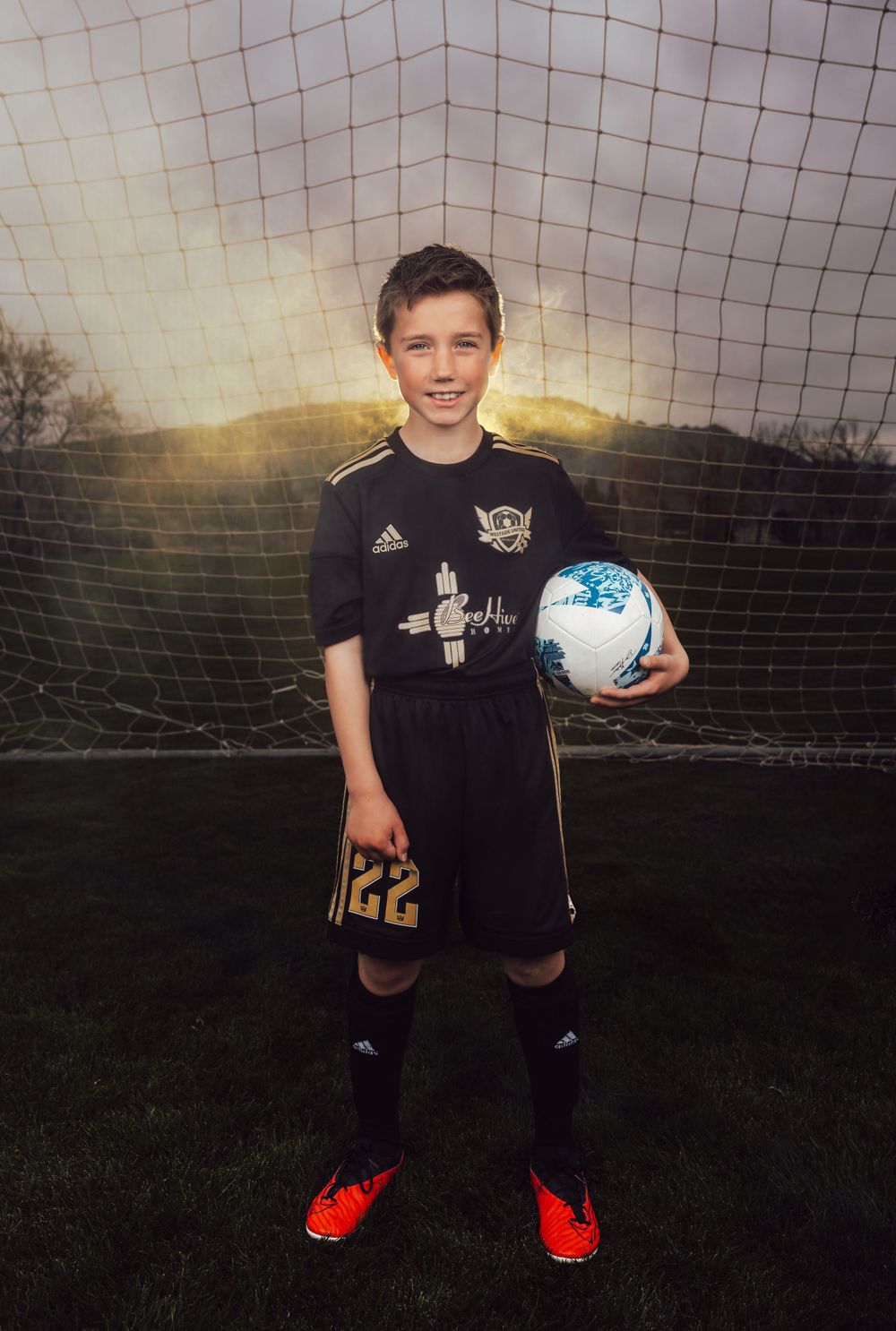 Soccer player holding blue ball poses in black uniform against sunset goal net.
