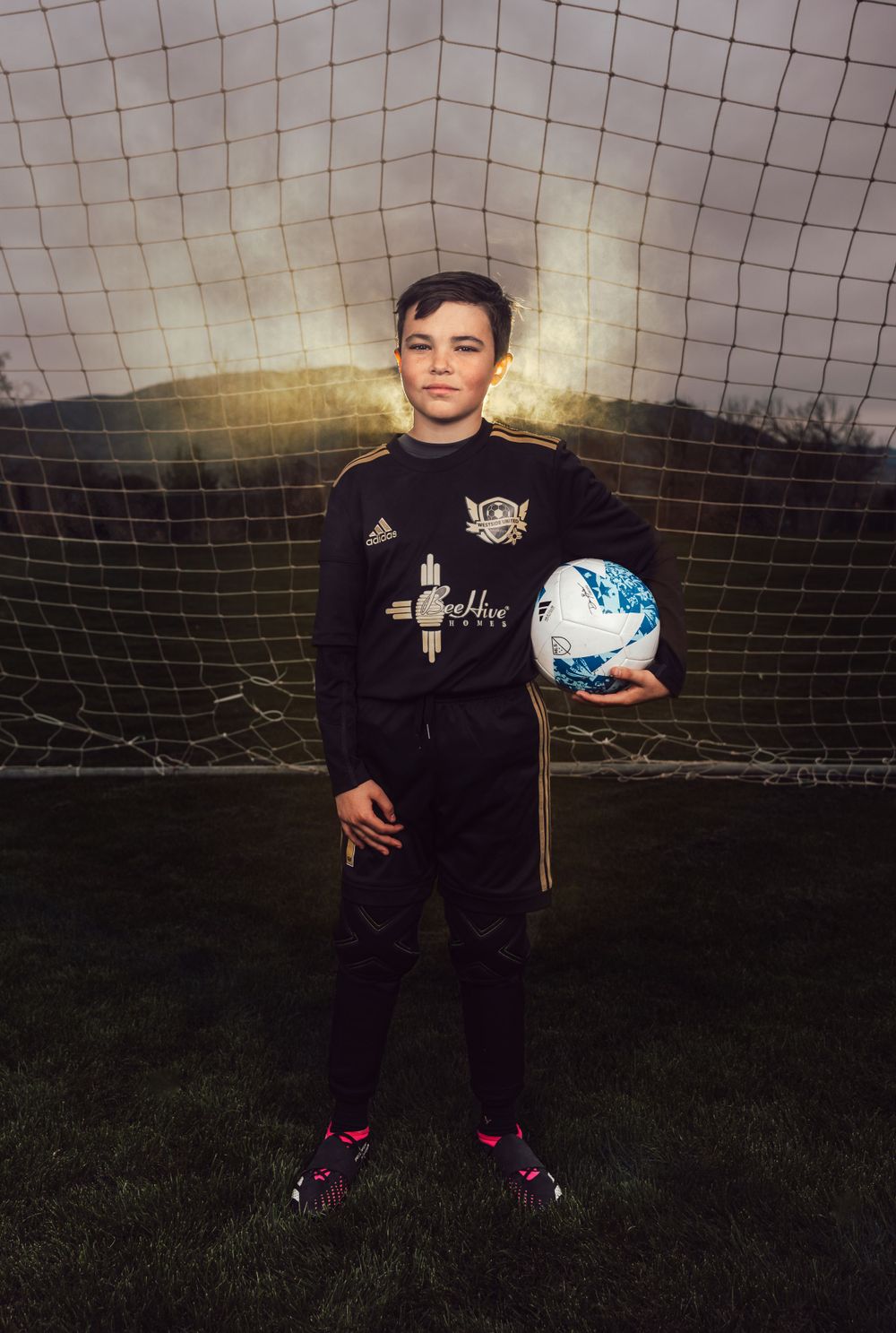 Soccer player holds blue and white ball while standing in front of goal net at dusk.