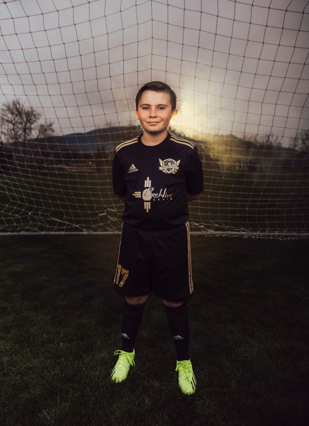 Player in black soccer uniform poses in front of goal net wearing neon cleats.