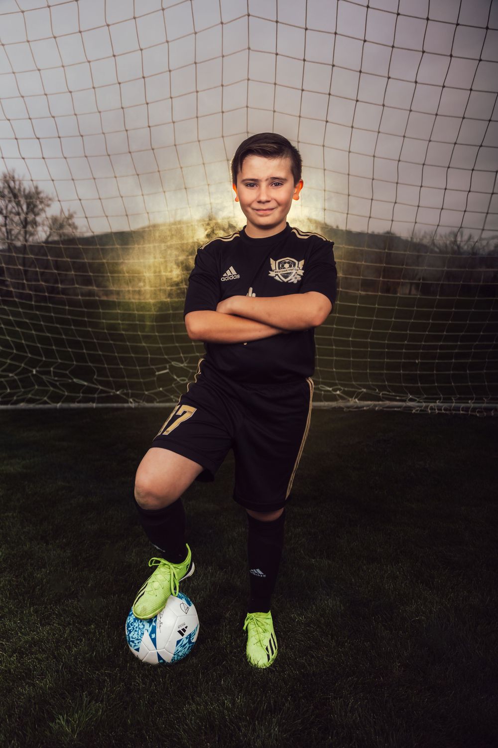 Soccer player in black uniform poses with ball at feet in front of goal net.