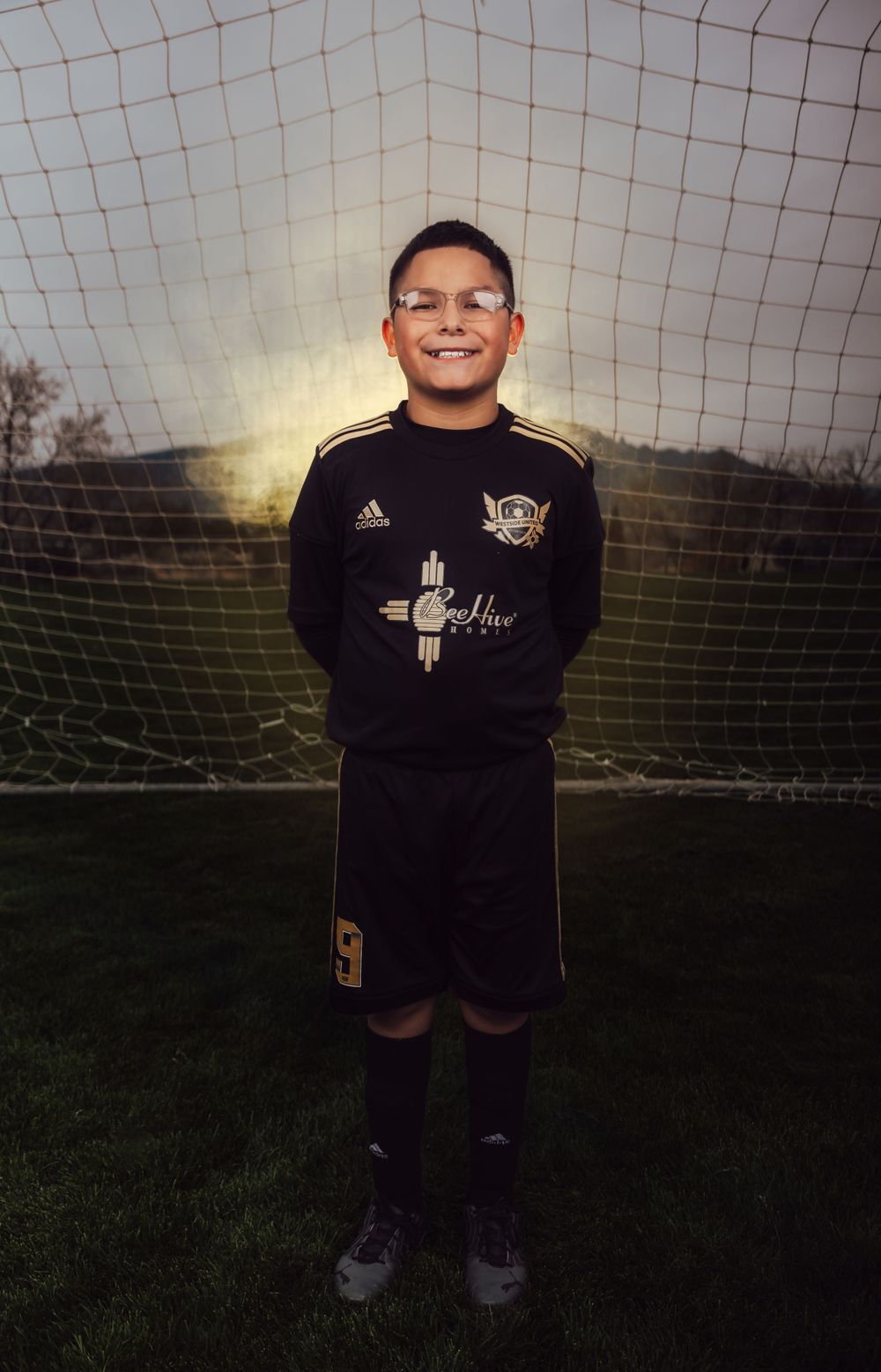 Soccer player in black Adidas uniform stands smiling against sunset goal net background.