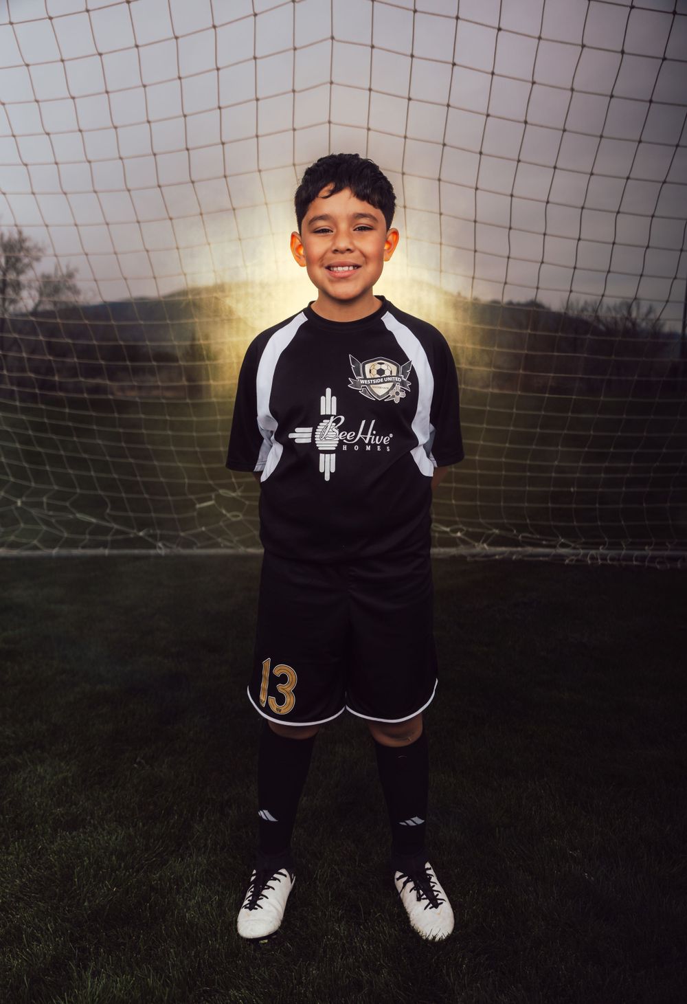 Youth soccer player in black uniform smiles in front of goal net during golden hour.