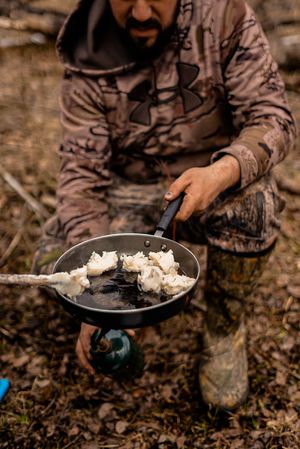 Person in camouflage clothing scooping flour mixture with a spoon in a forest setting.