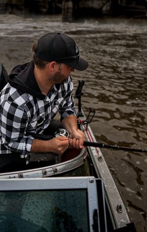 Person in plaid shirt sitting in a boat on murky water near a bridge.