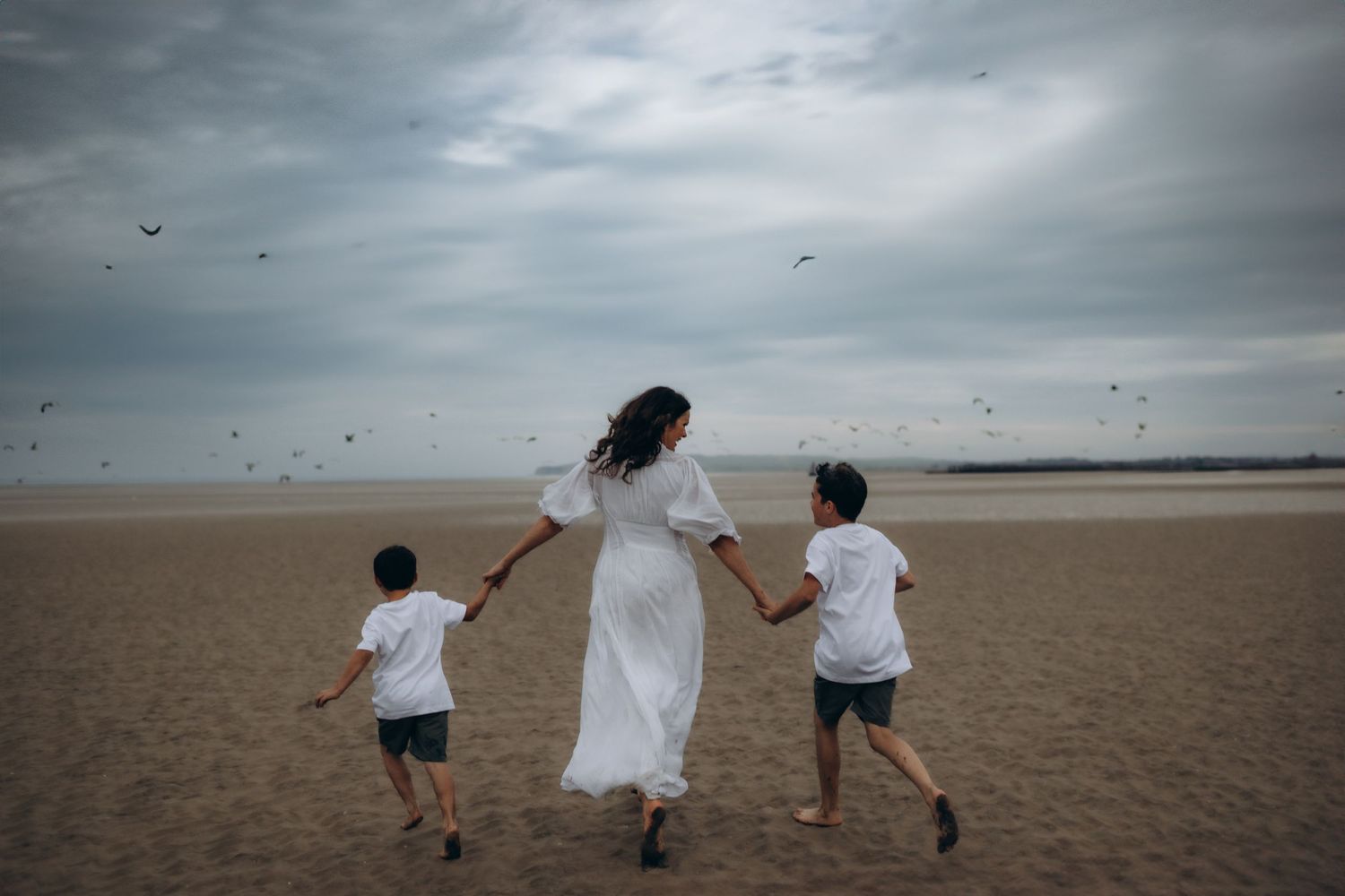 A mother holding hands with her two sons and running poetically towards the sea during their family photo session in Camber Sands, Kent
