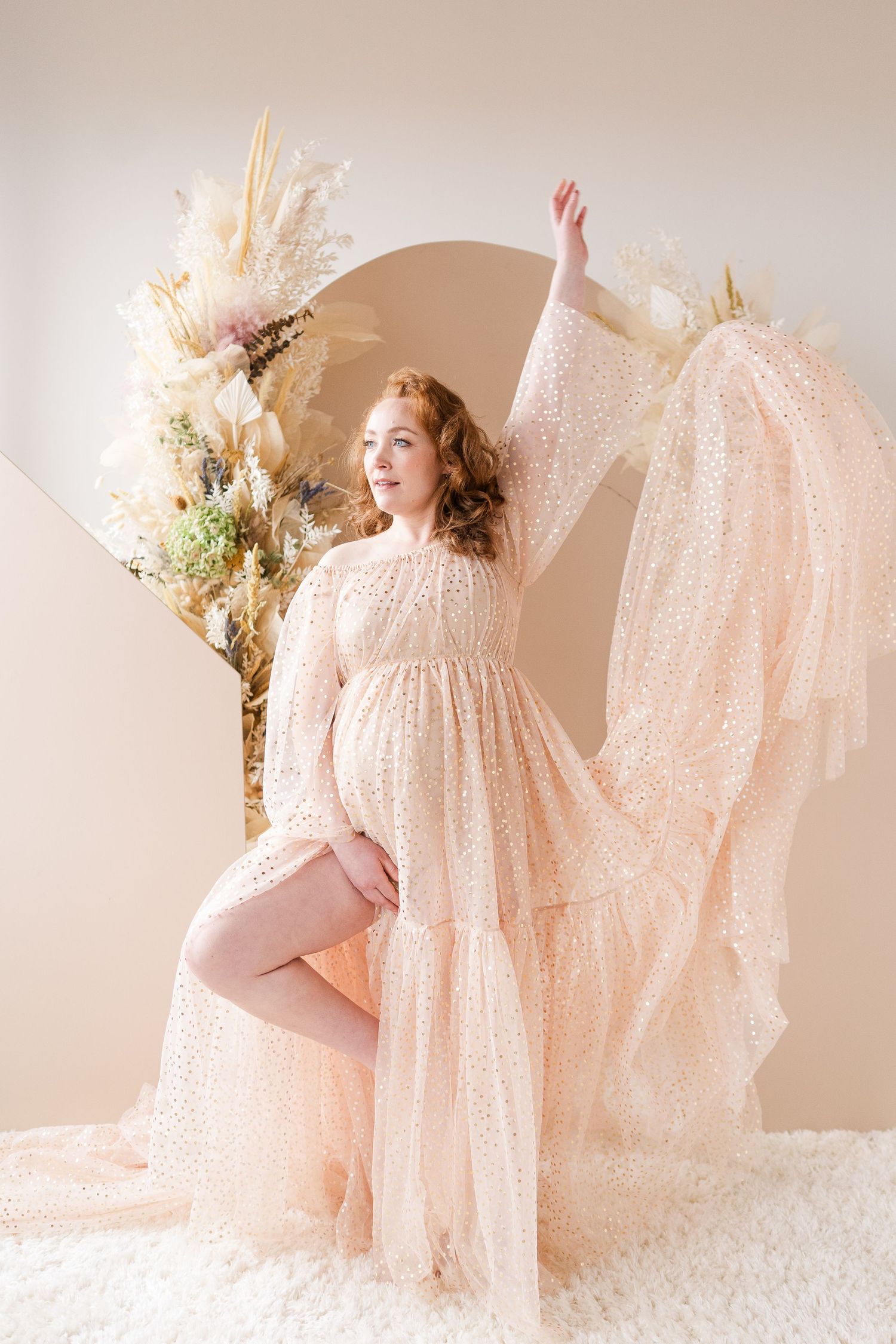 Whimsical maternity portrait in sparkly cream dress with artistic pose against arched backdrop.