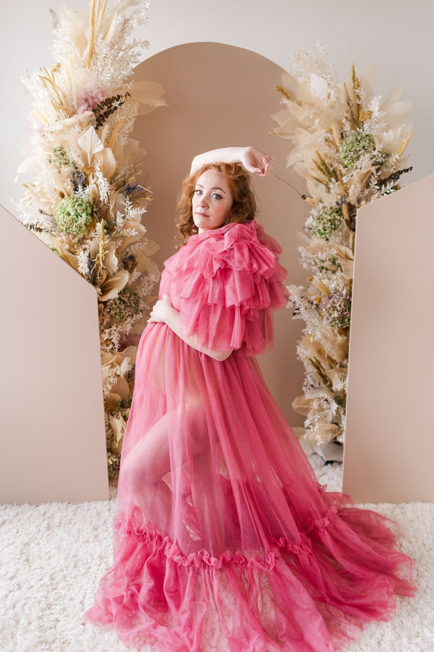 Glamorous maternity photo featuring flowing pink tulle gown with floral backdrop and dramatic styling.
