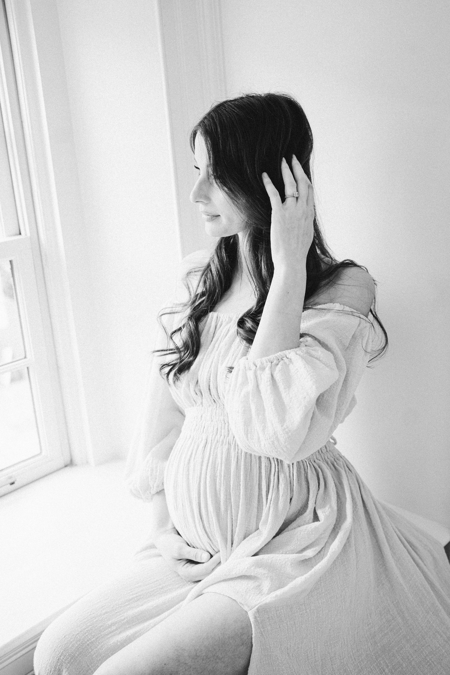 Black and white maternity portrait showing profile view near window in vintage style dress.
