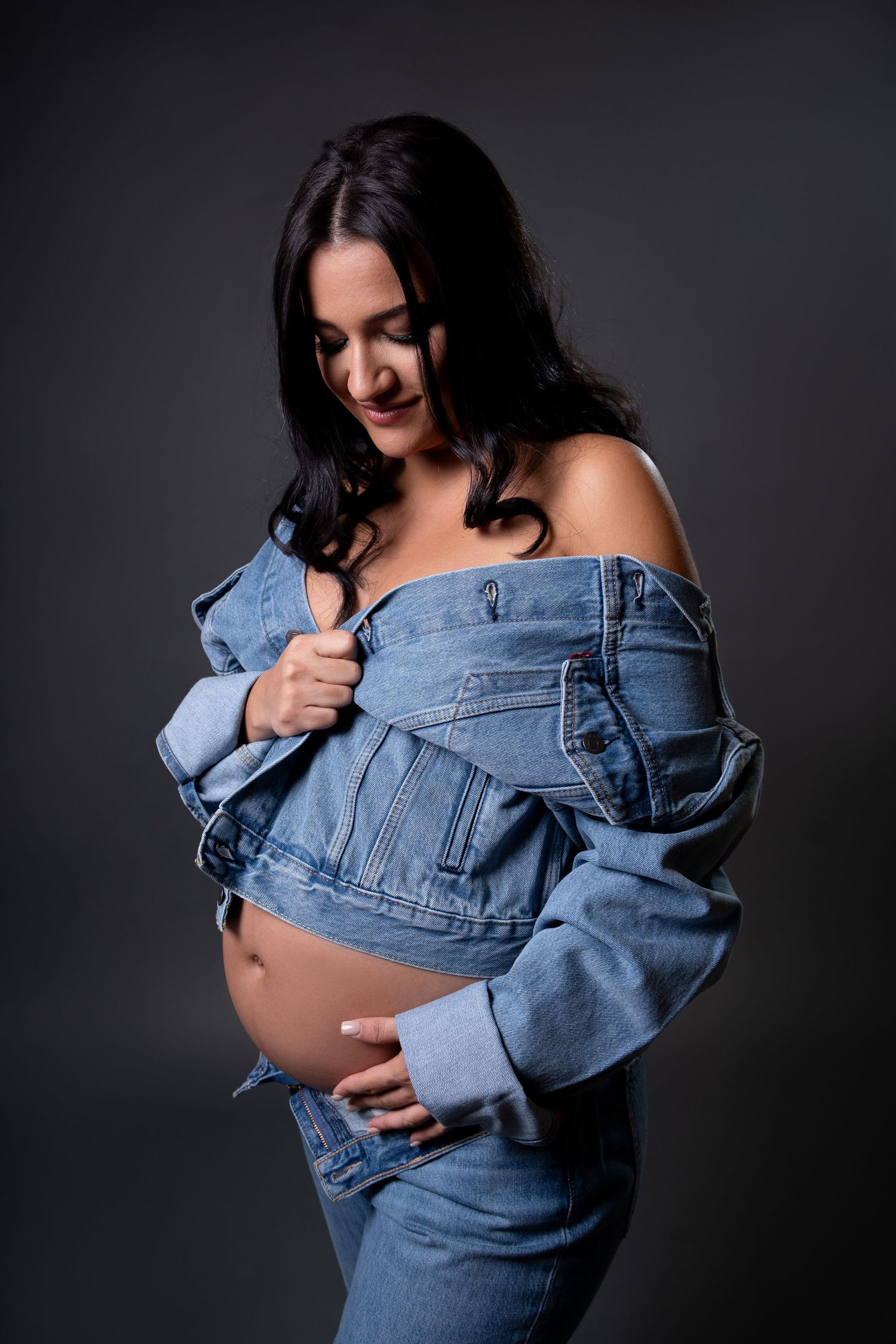 Creative maternity pose with denim jacket against dark backdrop.