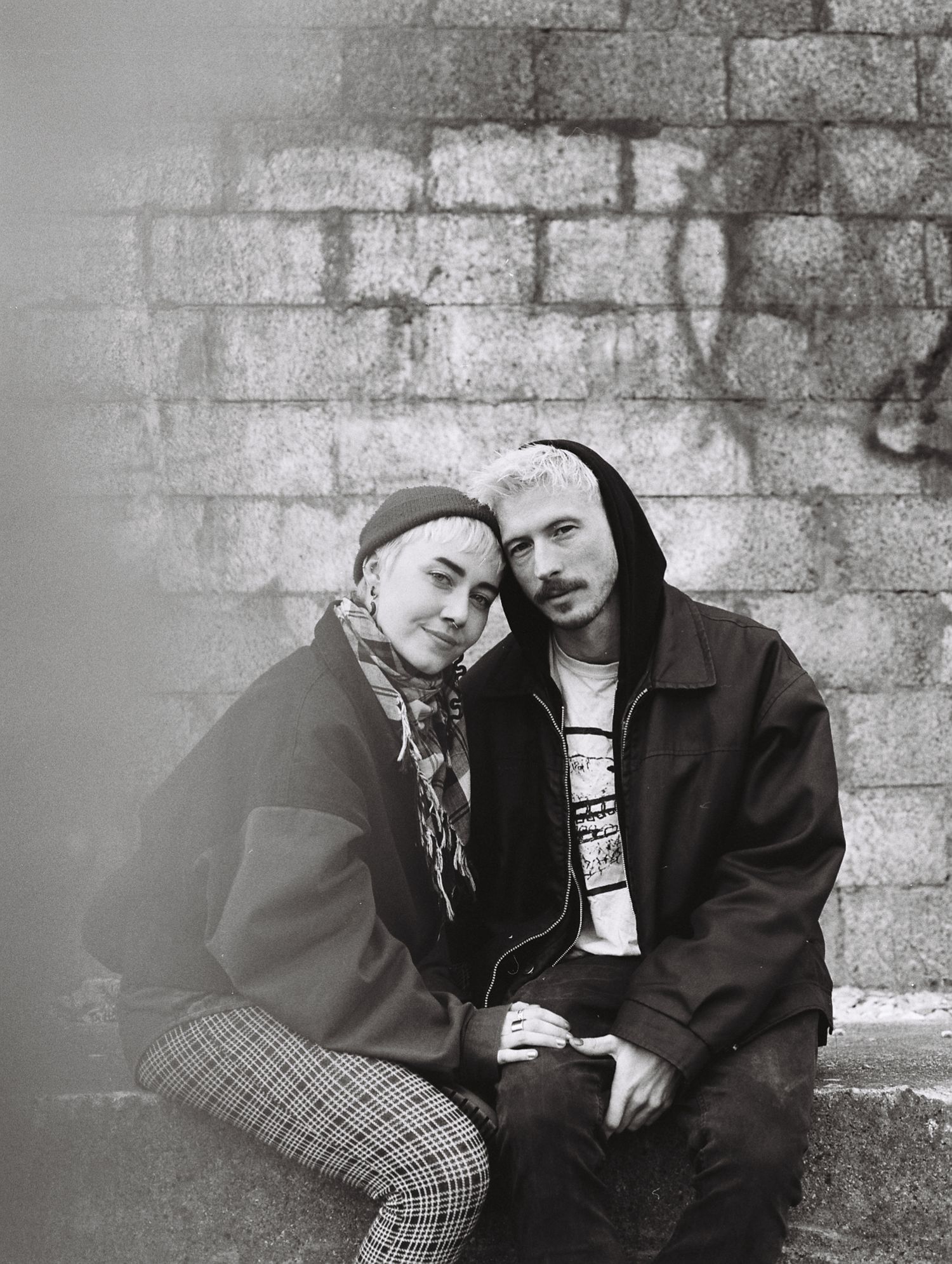 Intimate black and white portrait of a couple posed against a textured brick wall.