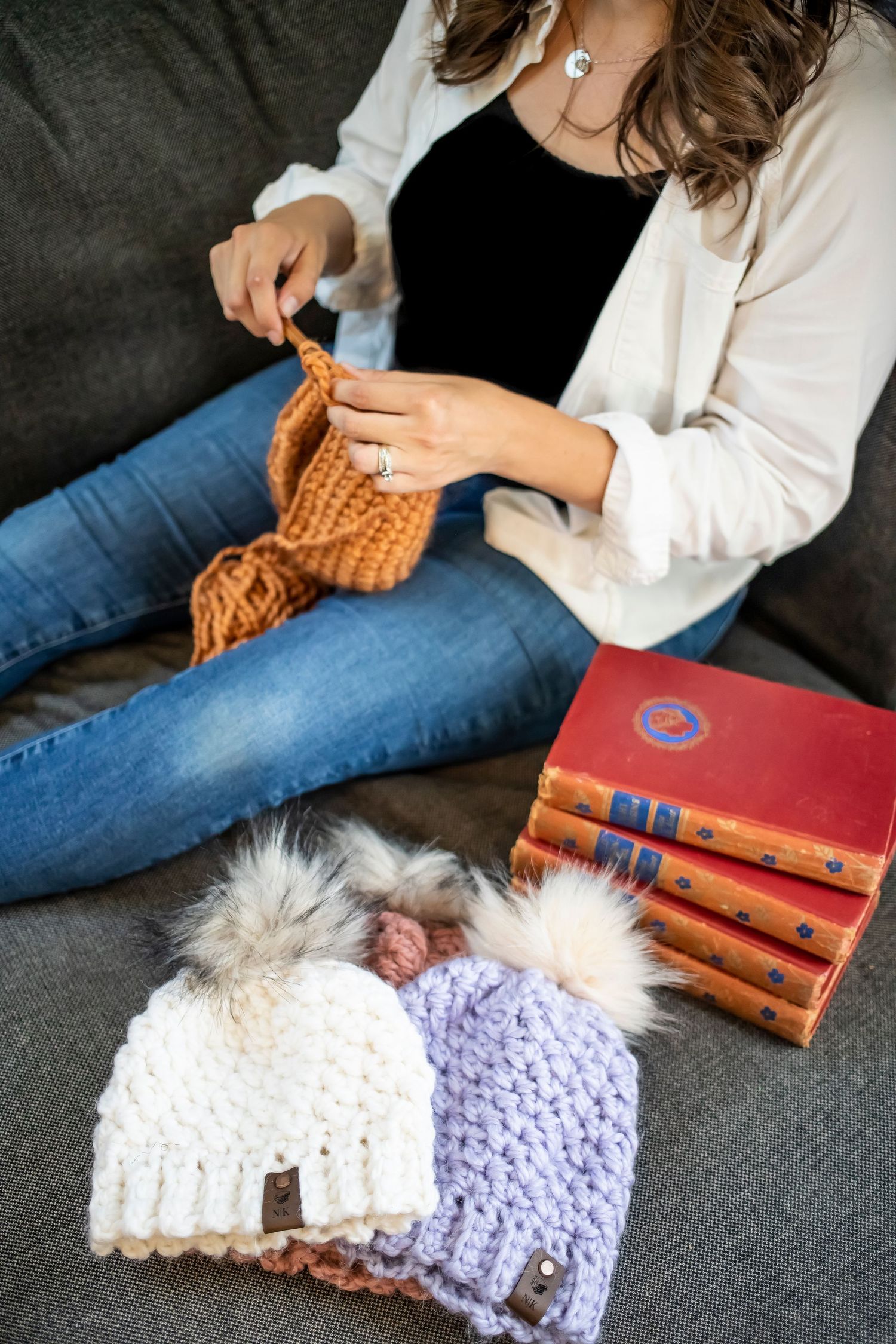 Person knitting with orange yarn while sitting on couch with knit beanies and books nearby.
