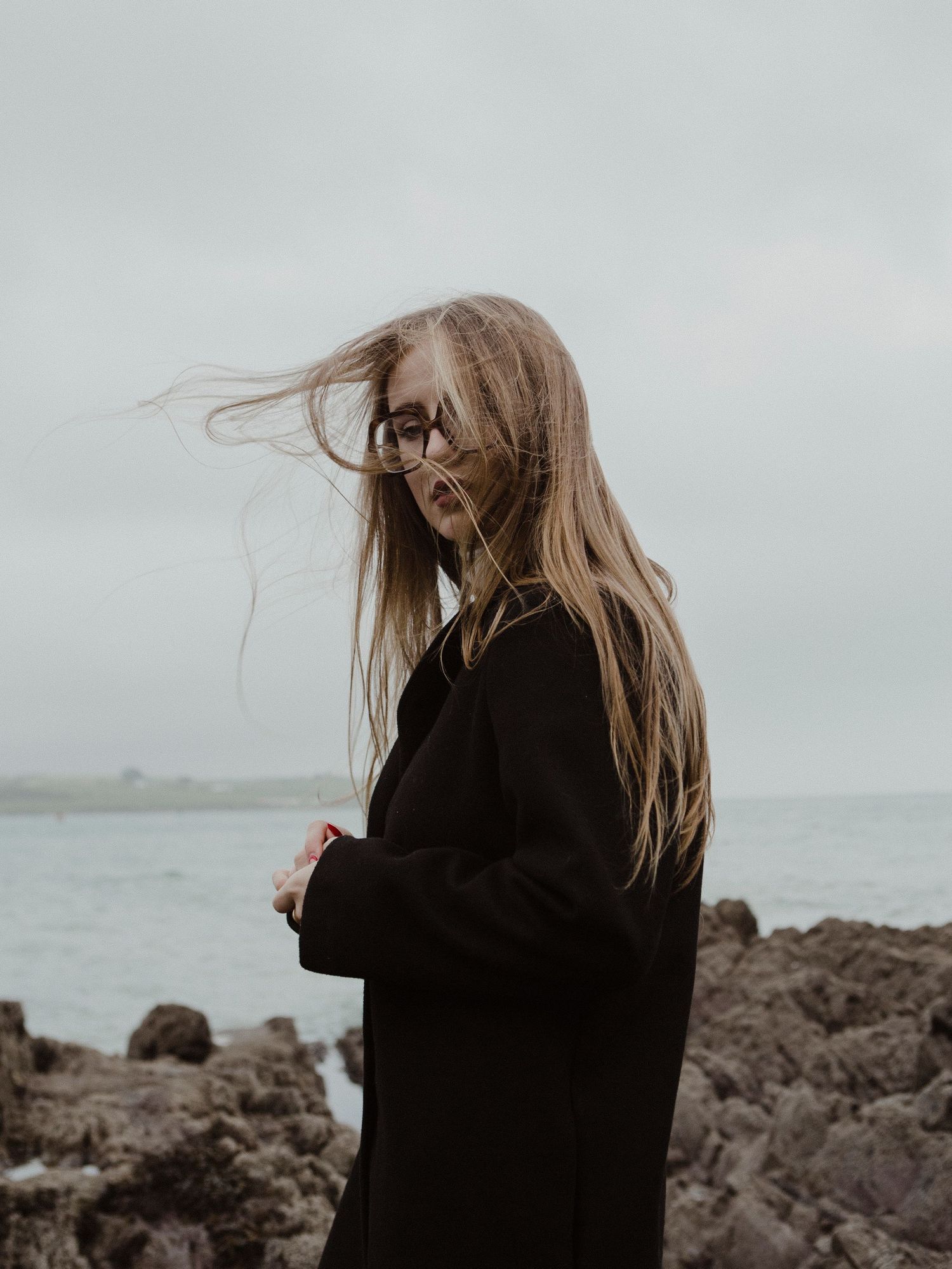 Moody portrait of a person in black coat standing on rocky shore with windswept hair.