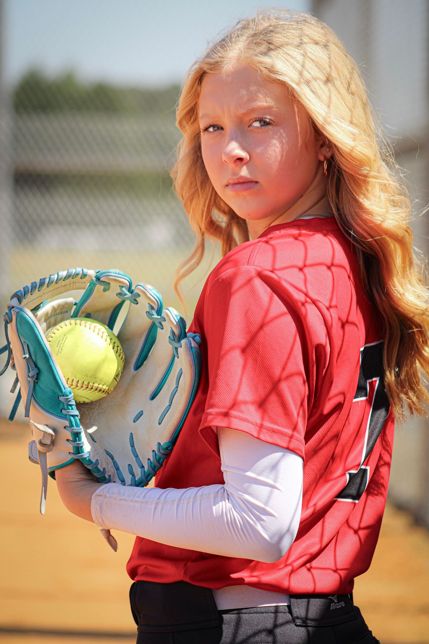 Softball player in coral jersey holding turquoise glove on baseball field.