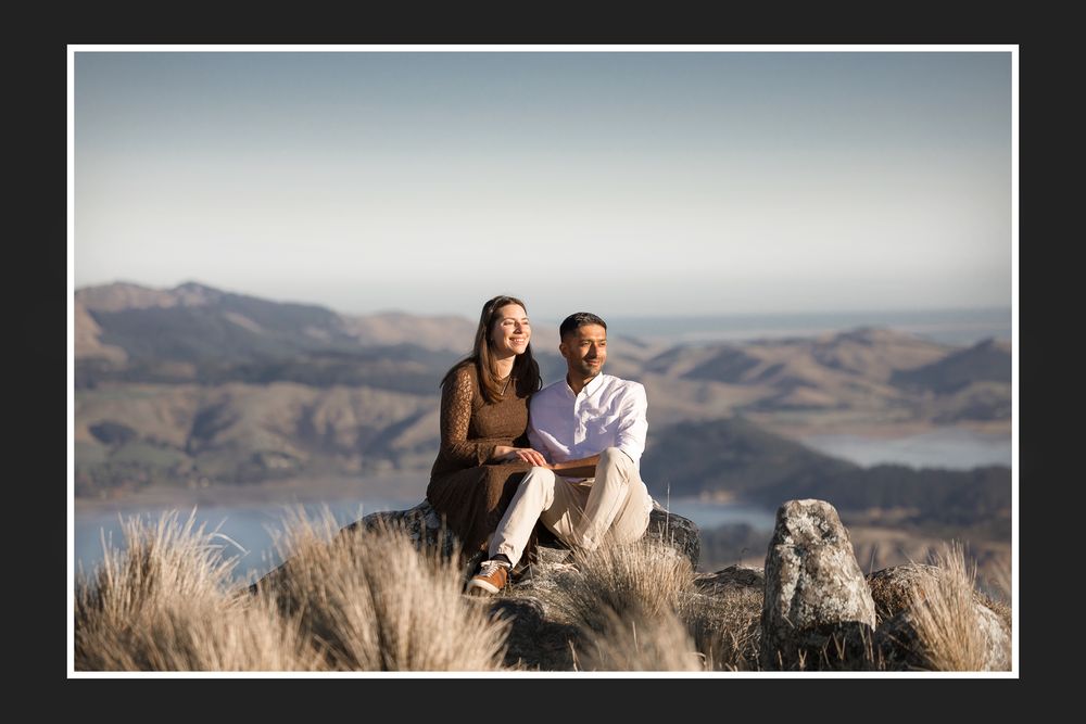 Couple sitting on rocky hilltop with scenic lake and mountain views in background on a clear day.