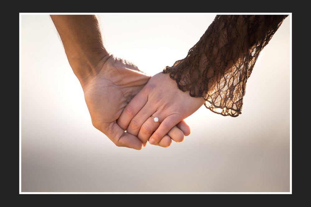 Close-up of a couple holding hands, woman wearing an engagement ring, with lace dress detail visible.