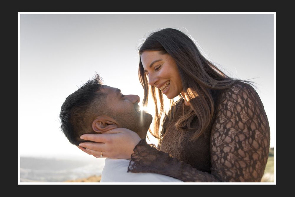 A couple shares a joyful, intimate moment outdoors, smiling at each other in warm backlit sunlight.