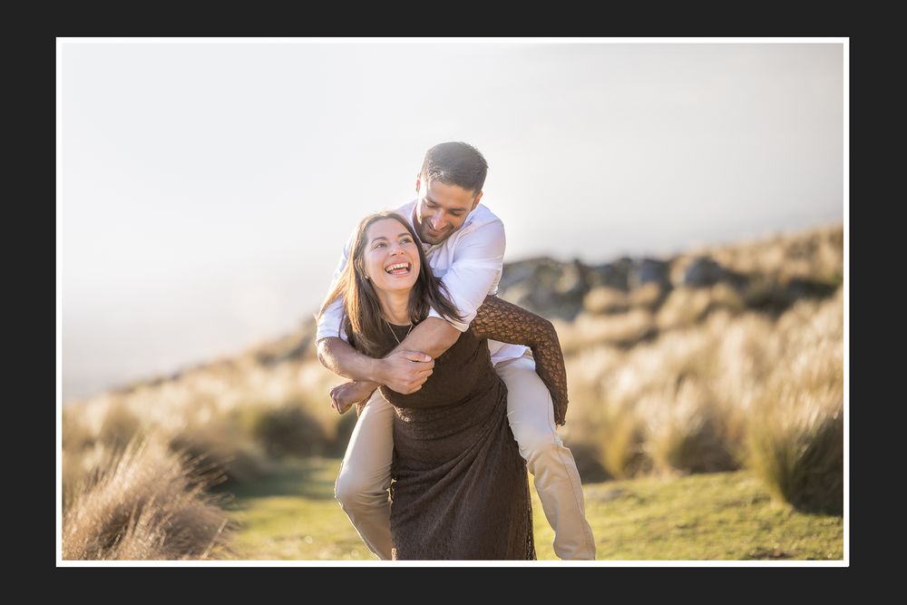 Couple laughing outdoors, woman giving man a piggyback ride in a sunny field during an engagement photo shoot.