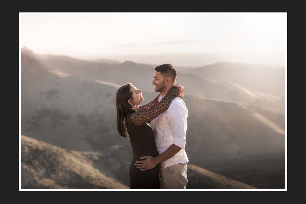 Couple embracing on a mountain overlook at golden hour with hazy hills in the background.