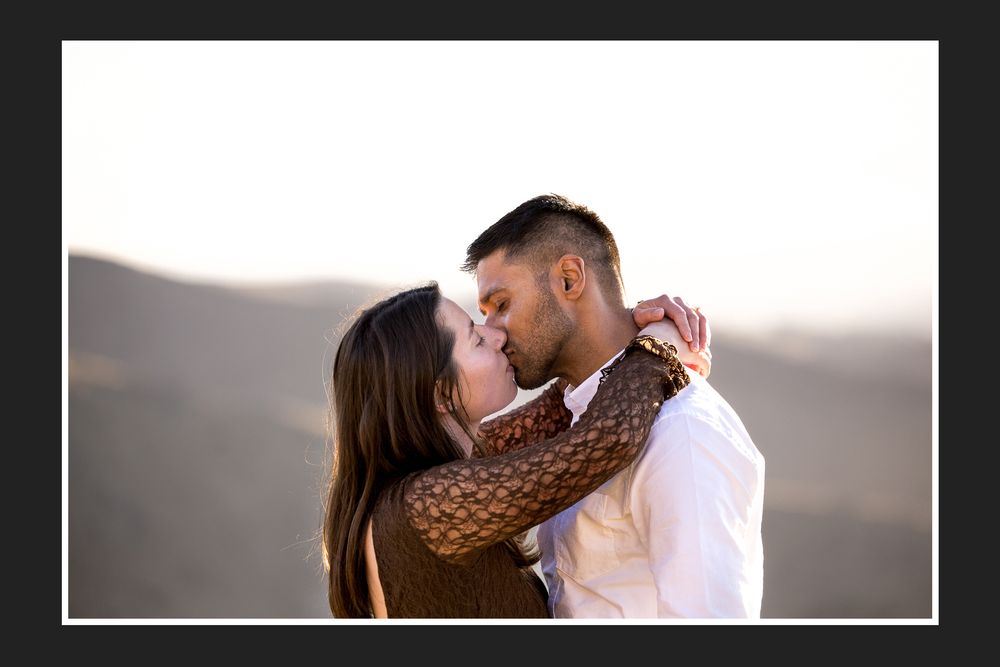 A couple shares a romantic kiss outdoors with scenic mountain landscape and golden sunset light in the background.