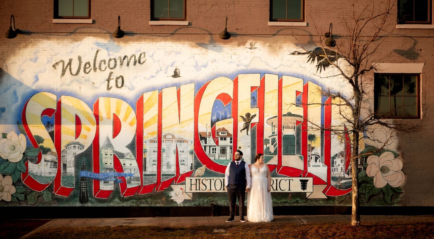 A colorful vintage-style 'Welcome to Springfield' mural painted on a brick wall with a wedding couple posing in front.